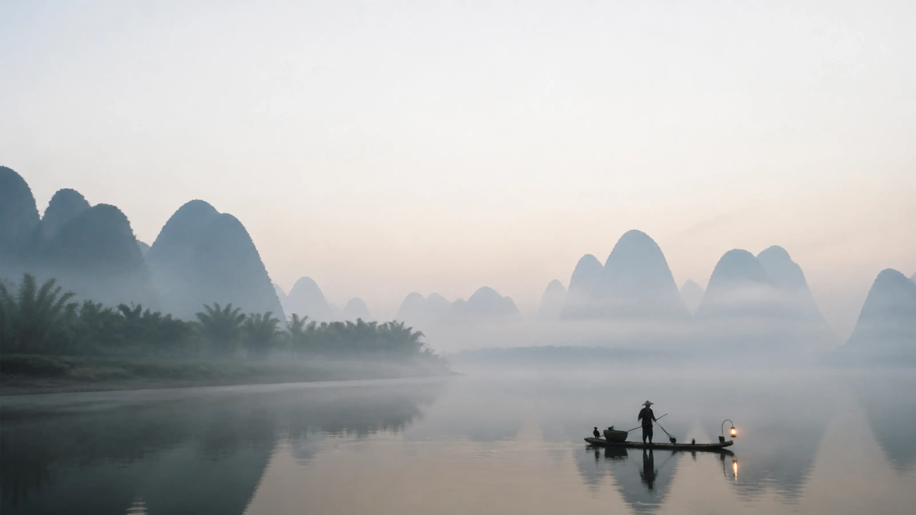 A fisherman poling a bamboo raft through morning mist on the Li River, karst peaks layered behind
